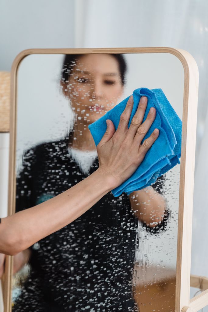 Close-up of a person wiping a mirror with a blue cloth, showing hands and reflection.